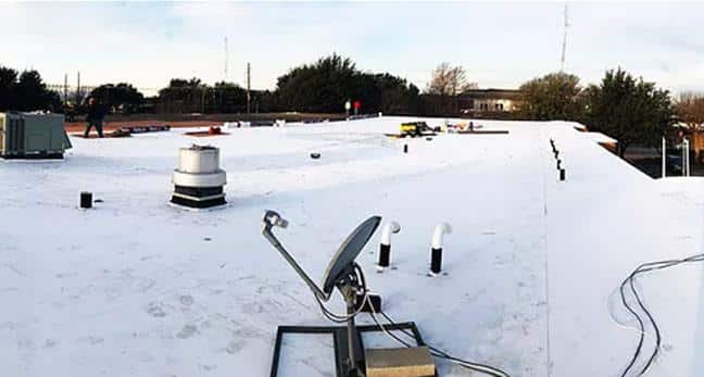 A wide view of a flat rooftop with a white surface. The roof has various equipment including a satellite dish, vents, pipes, and HVAC units. Several people are visible in the background near the edge of the roof. Trees and other buildings are seen in the distance.