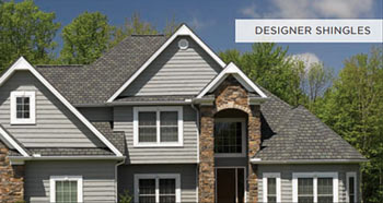 A large two-story house with gray siding and a complex roof featuring designer shingles. The home has multiple windows and a mix of stone and siding on the exterior. Trees are visible in the background, and a label reads "Designer Shingles.