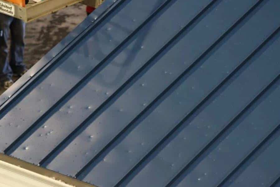 A close-up of a blue metal roof with visible dents. The roof has distinct, parallel grooves running vertically across it. Part of a person and some construction equipment are visible in the background on the left side.