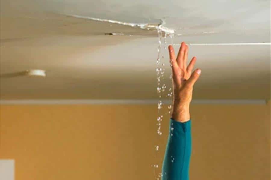 A hand reaches up to touch a ceiling with a visible crack, through which water is actively leaking. The scene suggests a situation where someone is attempting to handle a water leak emergency.
