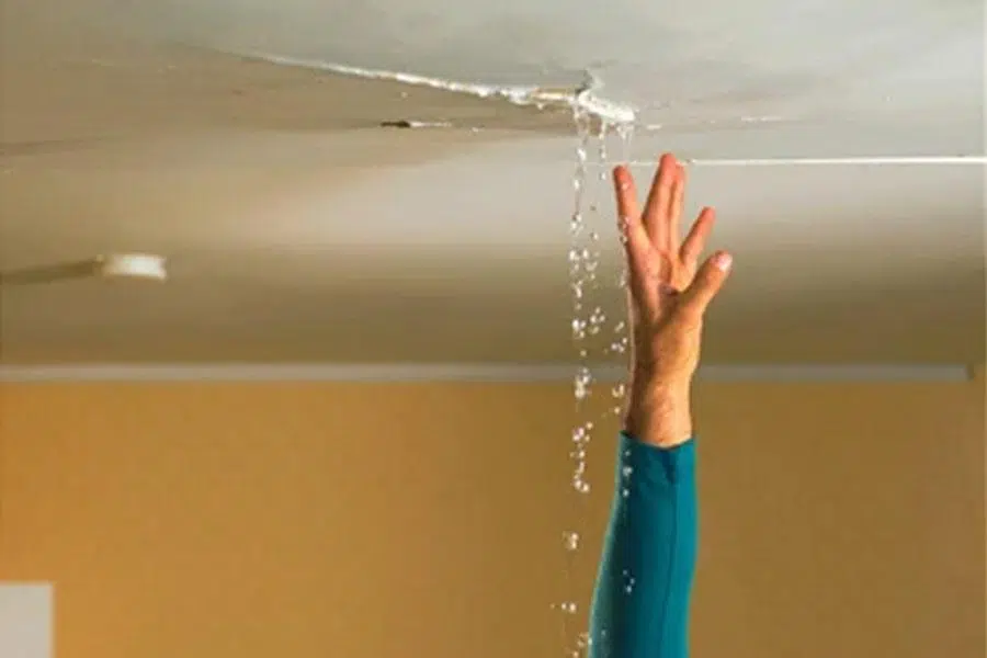 A hand reaches up to touch a ceiling with a visible crack, through which water is actively leaking. The scene suggests a situation where someone is attempting to handle a water leak emergency.