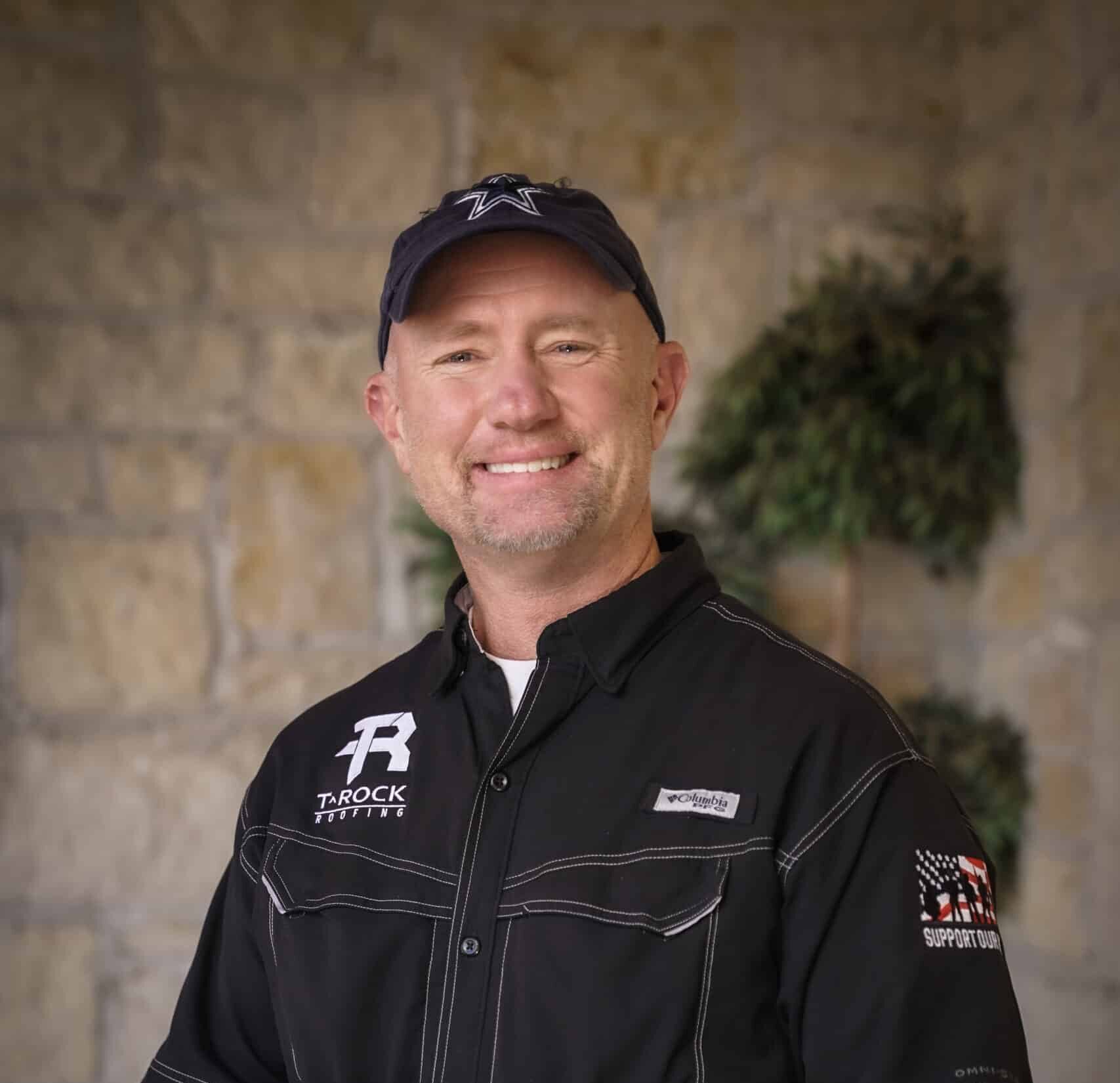 A smiling man wearing a black button-up shirt, a navy baseball cap, and blue jeans stands in front of a stone wall with greenery in the background.