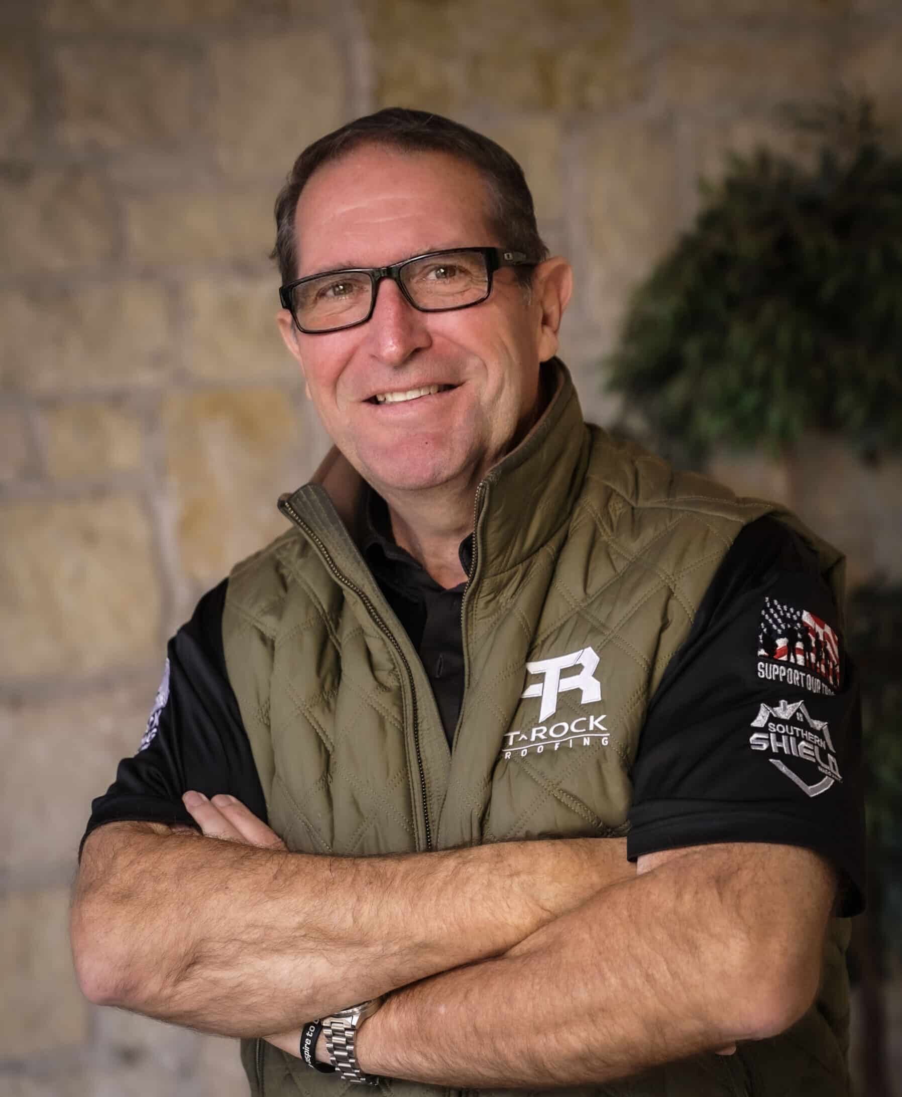 A man with glasses and short brown hair smiles with arms crossed. He wears a quilted green vest over a black polo shirt featuring logos and text. The background is a stone wall with a leafy plant to the side.