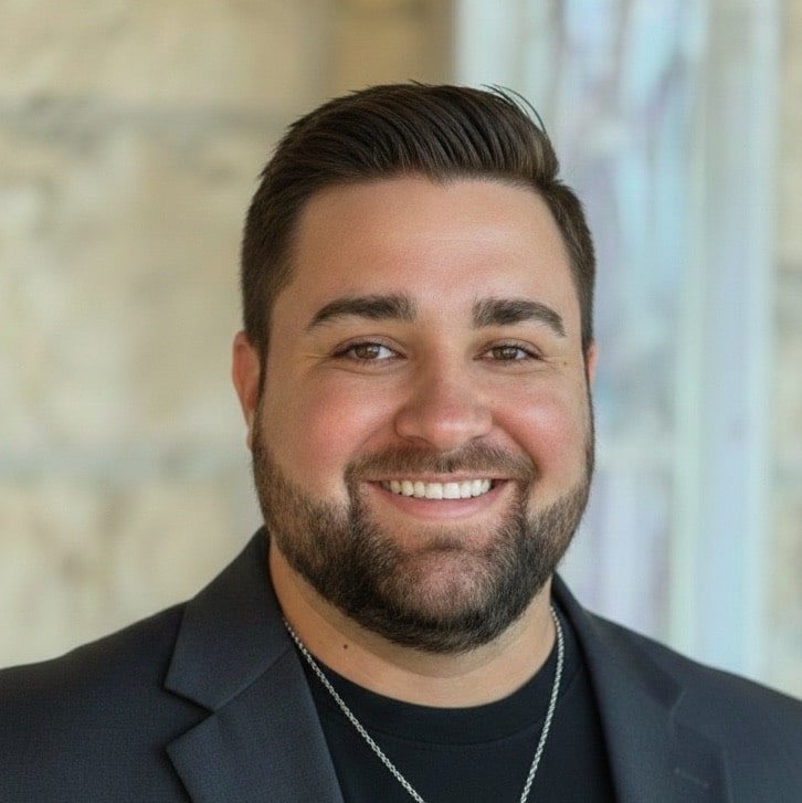 A man with short brown hair, a trimmed beard, and a friendly smile is wearing a black blazer, a black shirt, and a silver necklace, standing in front of a blurred light-colored background.