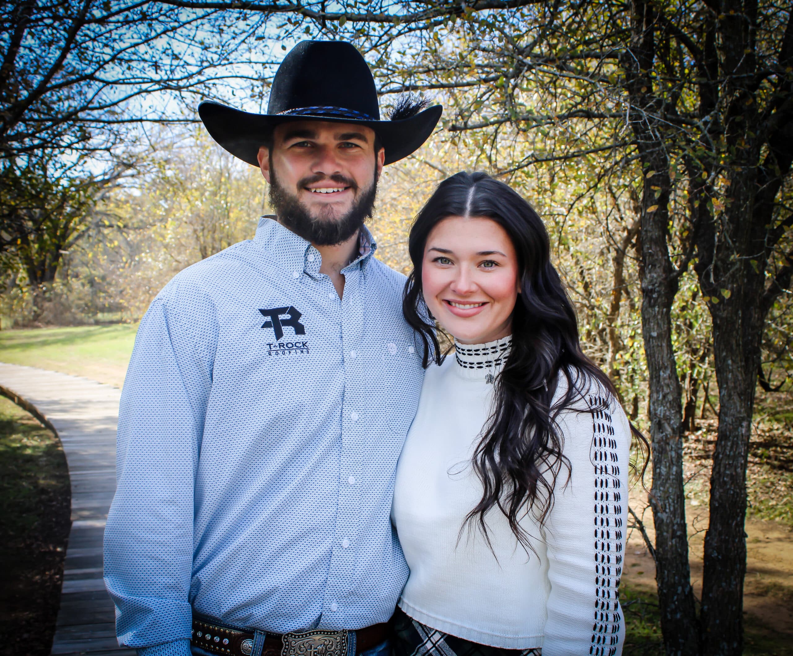 A couple poses outdoors on a wooden path surrounded by trees. The man wears a cowboy hat, a light blue shirt, and jeans. The woman wears a white sweater and a plaid skirt. Both are smiling, with the sun filtering through the branches. - T Rock Roofing and Contracting A couple poses outdoors on a wooden path surrounded by trees. The man wears a cowboy hat, a light blue shirt, and jeans. The woman wears a white sweater and a plaid skirt. Both are smiling, with the sun filtering through the branches.
