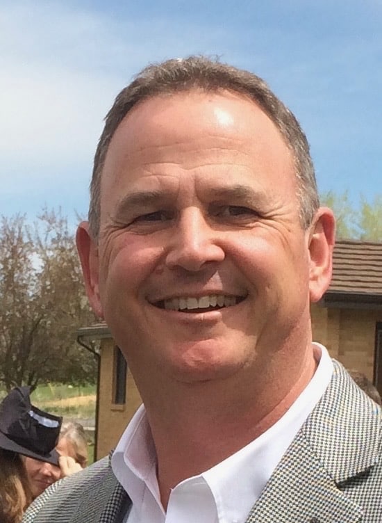 A middle-aged man in a gray checked blazer and white shirt smiles outdoors on a sunny day, with trees and a house in the background.