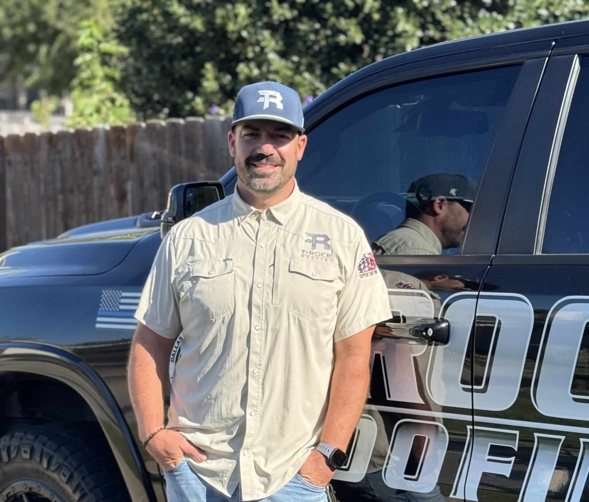 A man wearing a beige button-up shirt, blue jeans, and a black cap stands smiling by a black truck with "ROOF" lettering visible on the door. Trees and a wooden fence are in the background.