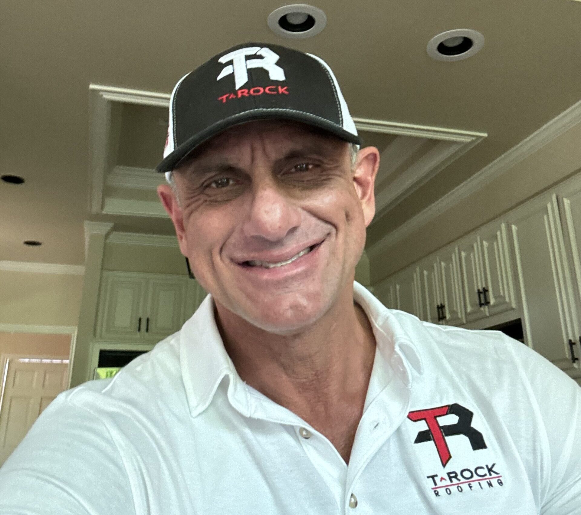 A man wearing a white polo and a black cap with the T-Rock logo smiles at the camera while standing in a kitchen with white cabinets and recessed lighting.