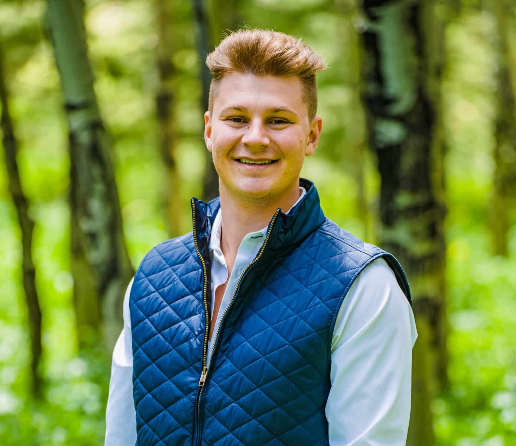 A young man stands outdoors in a forest, smiling. He wears a blue quilted vest over a white shirt and beige pants, with his hands in his pockets. Sunlight filters through the trees in the lush, green background.