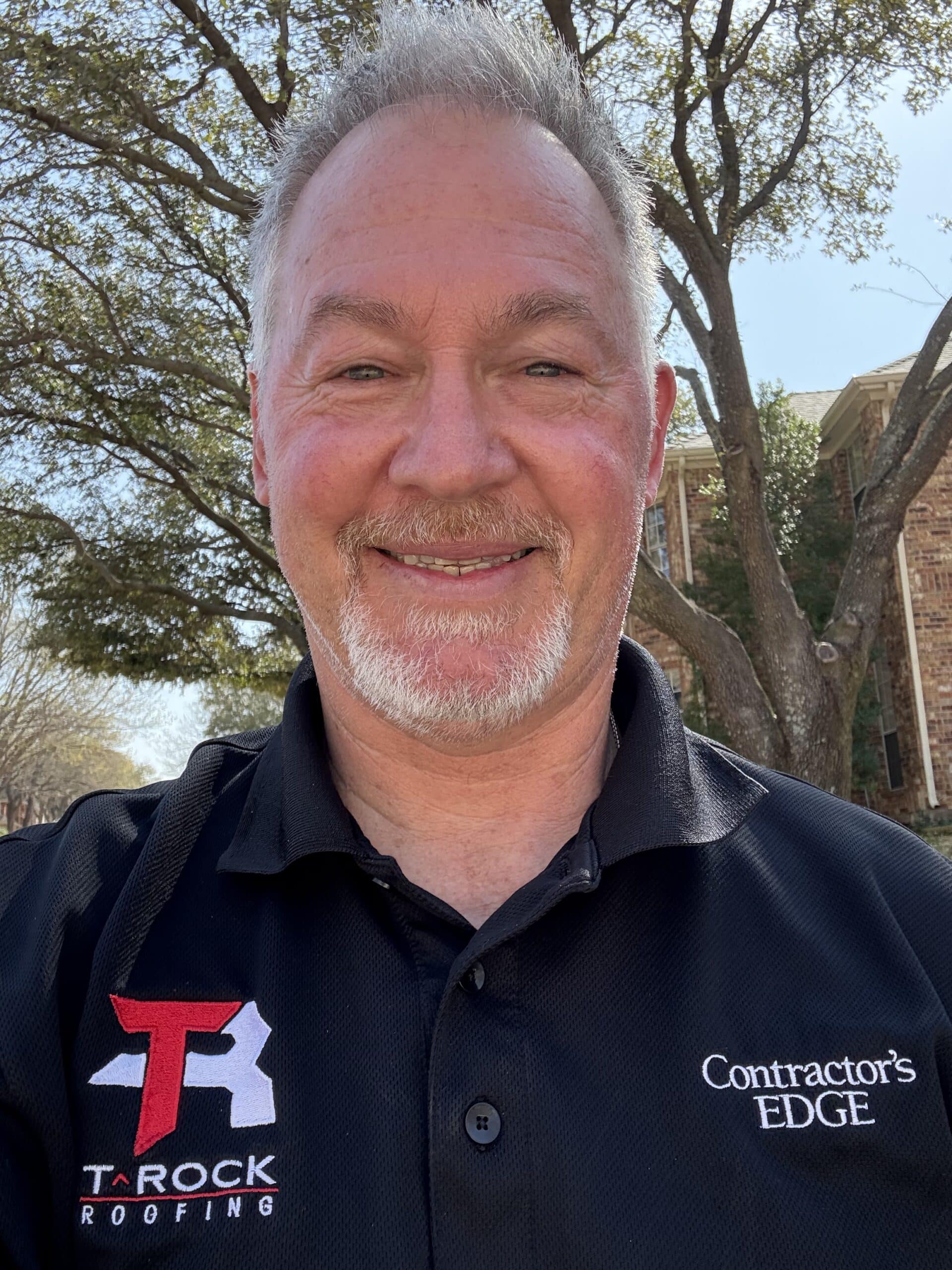 A man with short gray hair and a beard is smiling. He is wearing a black shirt with logos that read "Tarock Roofing" and "Contractor's Edge." Trees and a brick building are in the background.