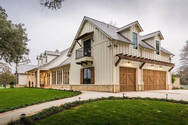 A modern farmhouse-style home with beige siding, large windows, and a metal roof. The house features a two-car garage with wooden doors and small balcony windows. The front yard is well-maintained with green grass and a paved driveway. Trees surround the property.