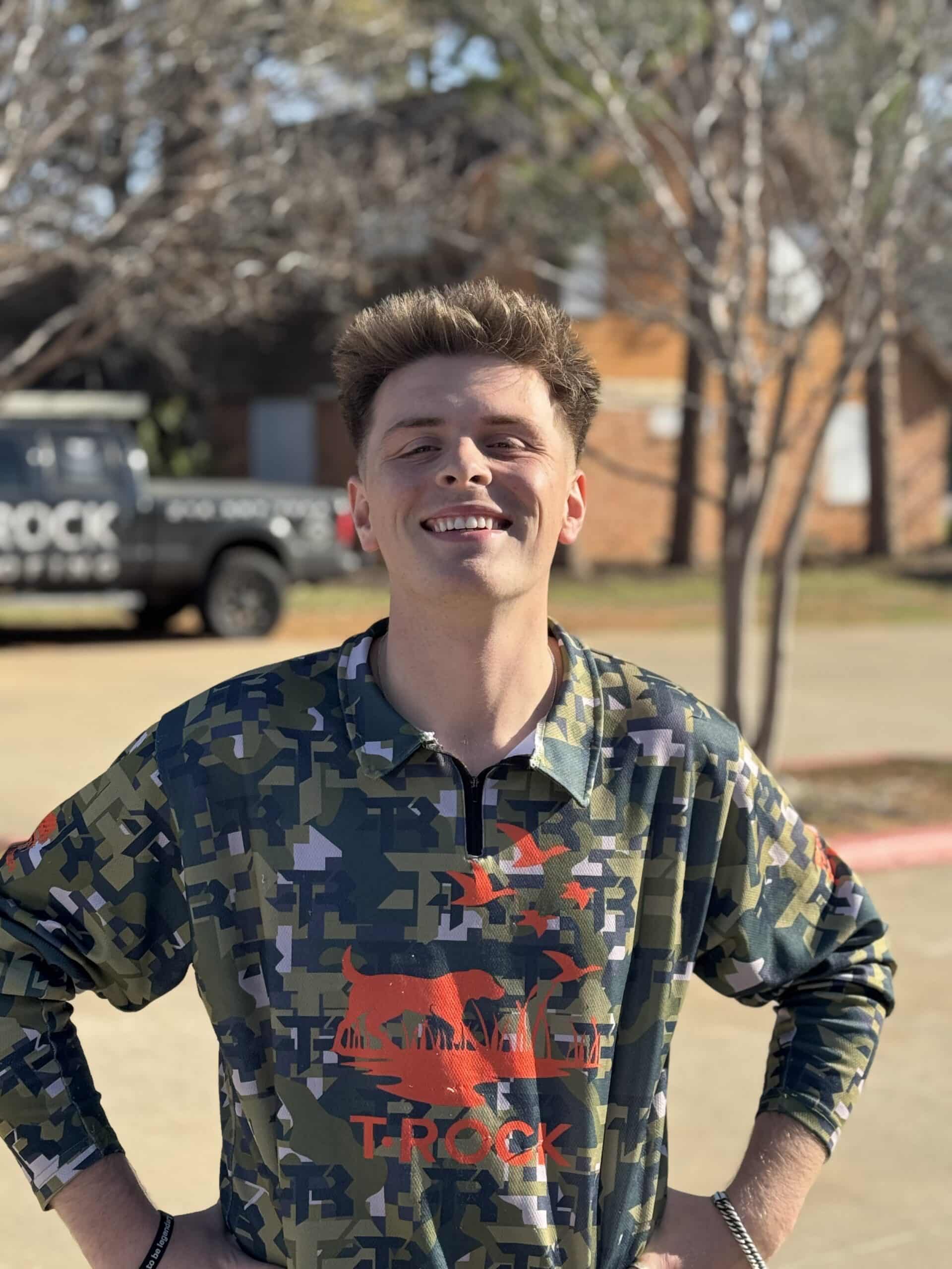 A young man with short brown hair smiles outdoors, standing with his hands on his hips. He wears a green and camo-patterned shirt with a red bear and the word "TROCK." Trees and a vehicle are visible in the background.