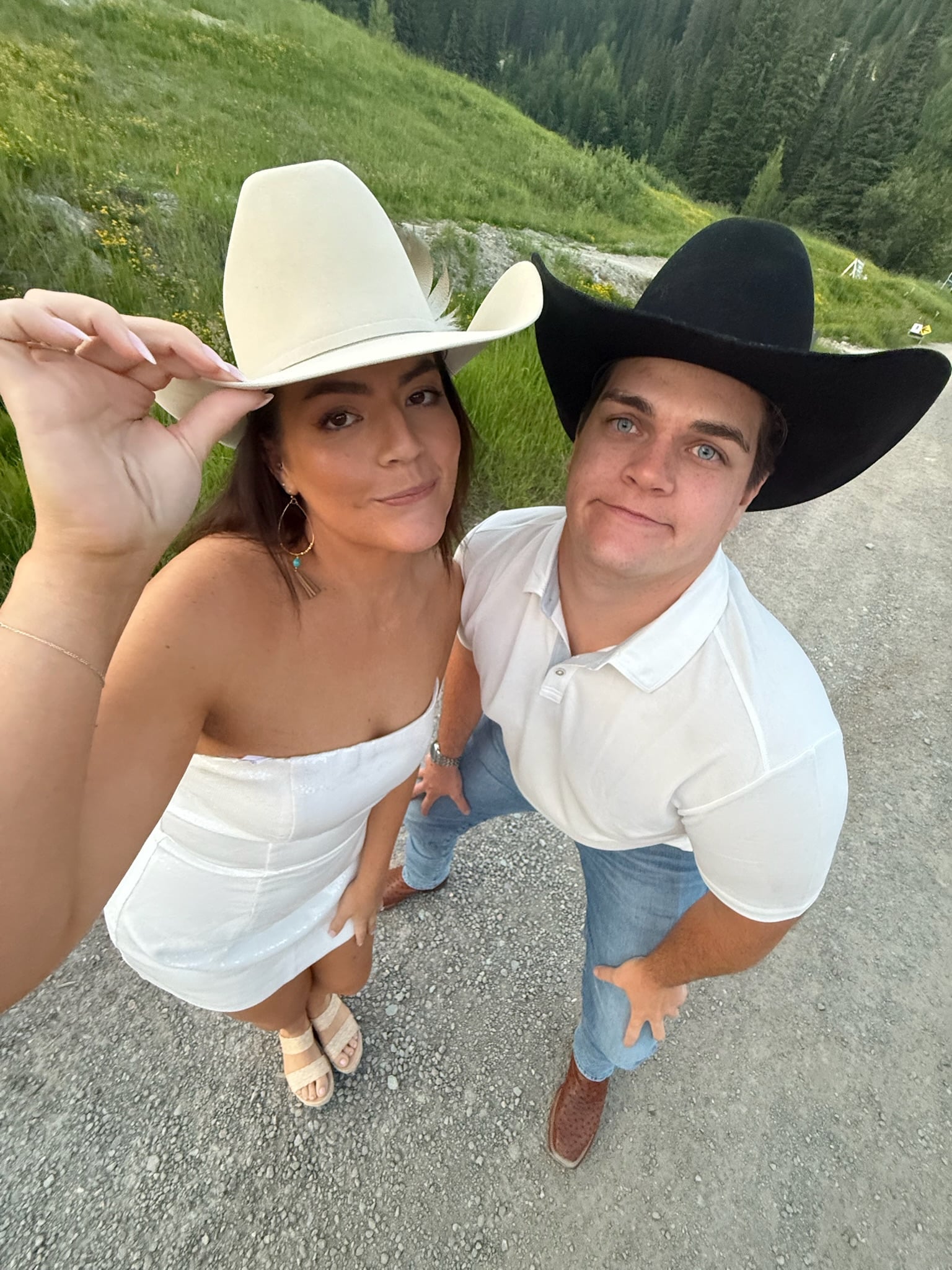 A woman in a white dress and hat stands next to a man in a white shirt and black hat on a gravel path with grass and trees in the background, both smiling and looking up at the camera.