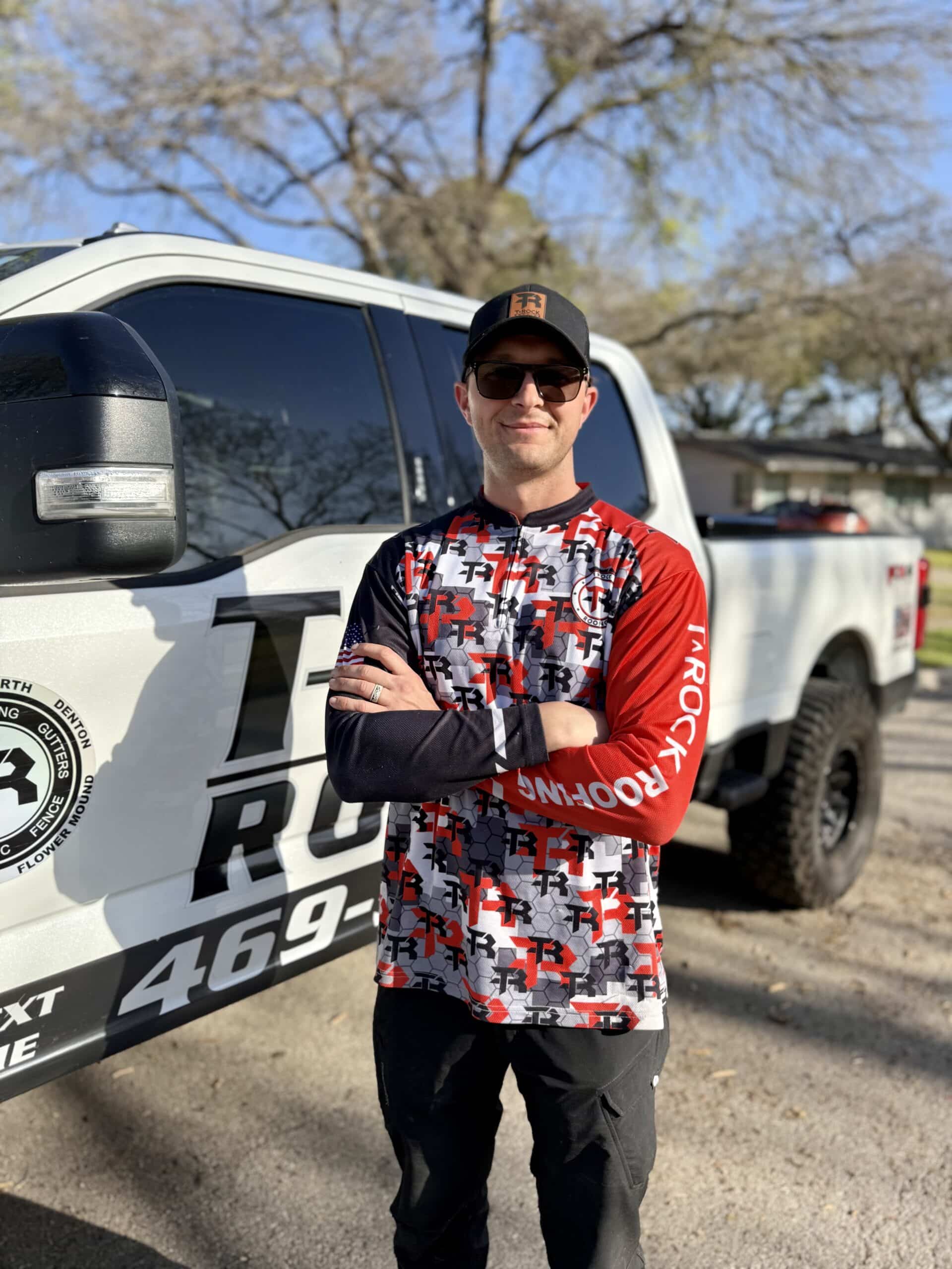 A man wearing sunglasses, a black cap, and a red, black, and white graphic shirt stands with arms crossed in front of a white pickup truck with business logos and large text on the door. Trees and sunlight are in the background. - T Rock Roofing and Contracting A man wearing sunglasses, a black cap, and a red, black, and white graphic shirt stands with arms crossed in front of a white pickup truck with business logos and large text on the door. Trees and sunlight are in the background.