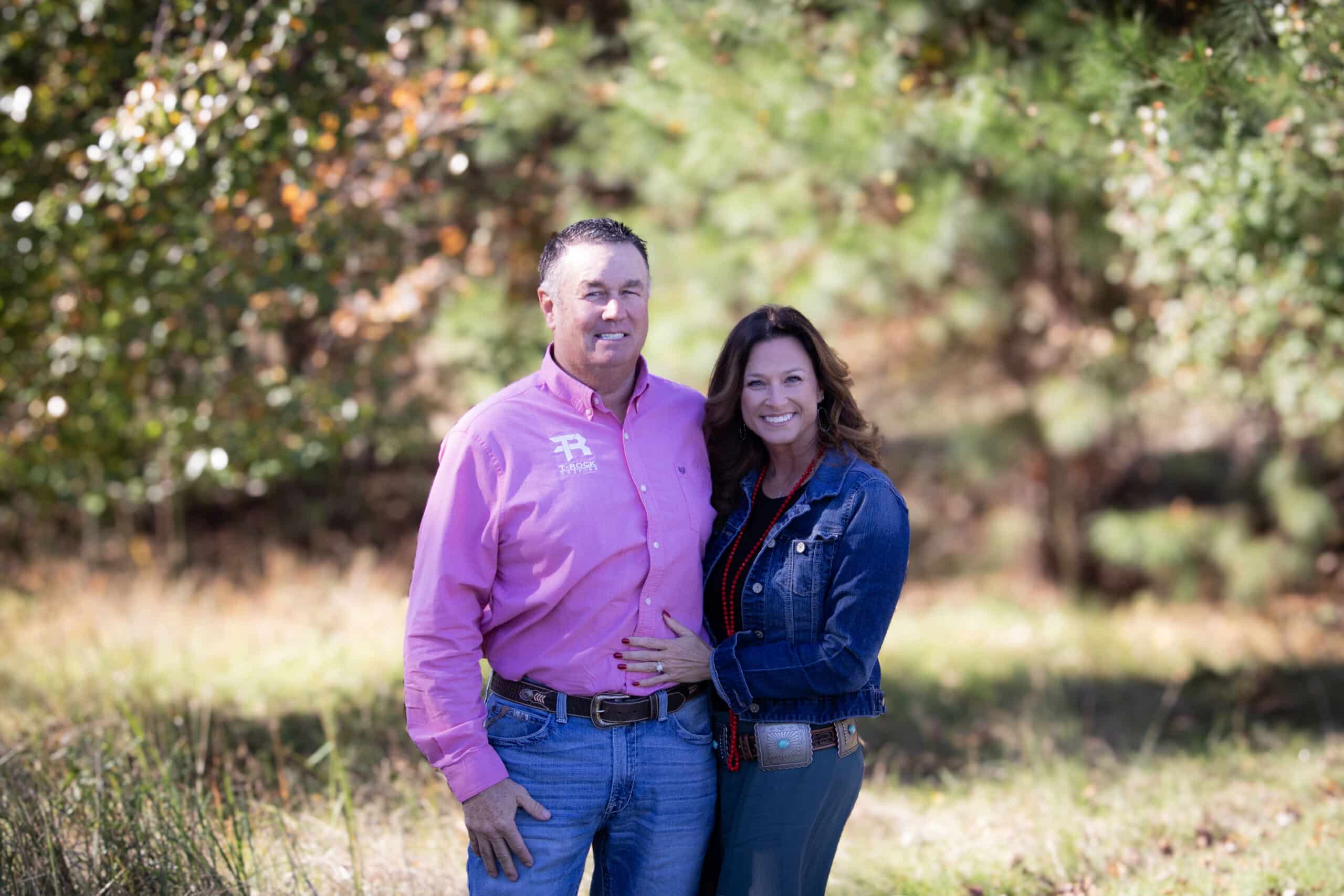 A man in a pink shirt and jeans stands beside a woman in a denim jacket and jeans. They are smiling in a sunlit outdoor setting with trees and grass in the background.