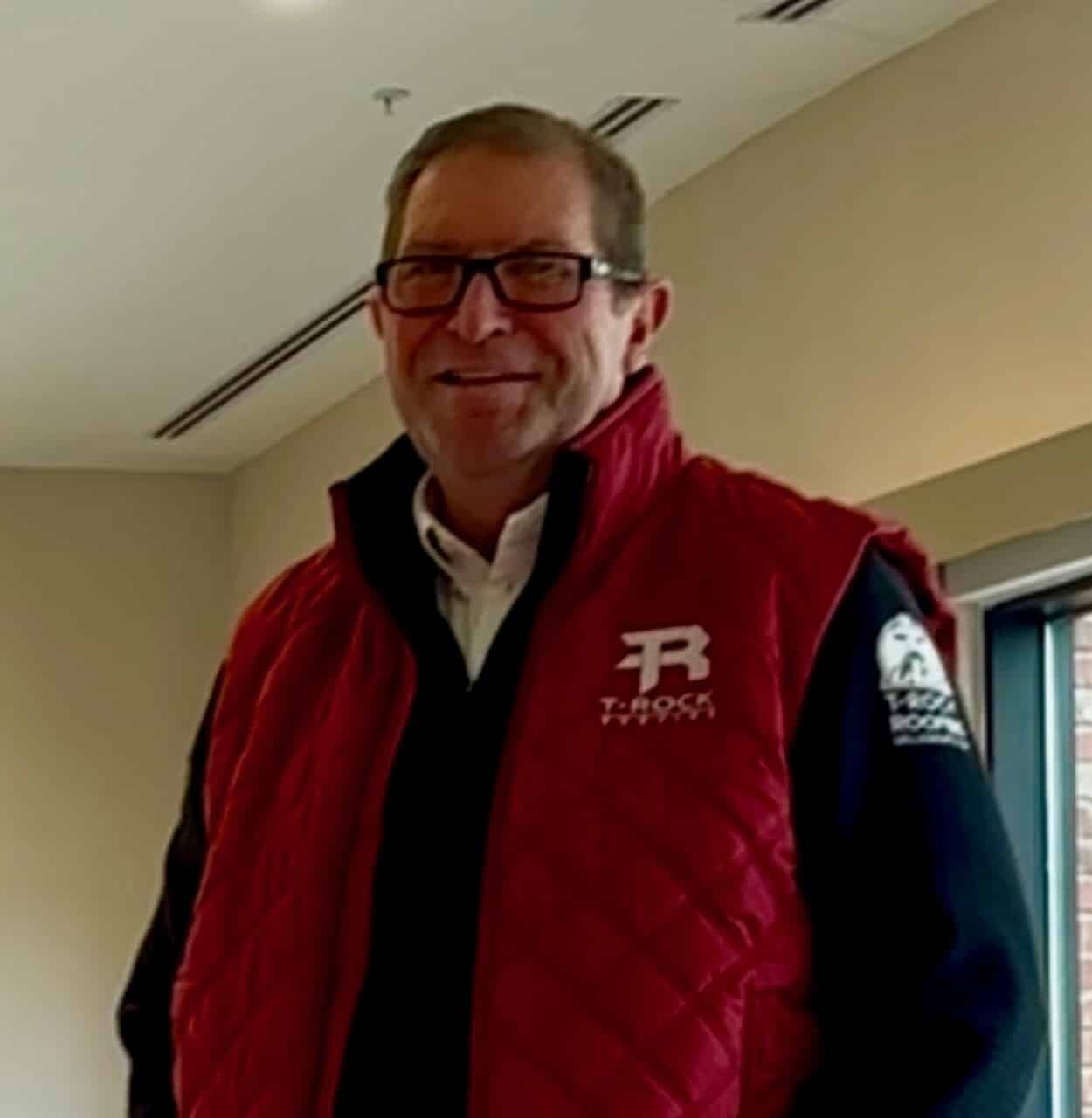 A man wearing glasses, a red quilted vest, and blue jeans stands indoors, smiling with one hand in his pocket. Light from a nearby window illuminates the scene.