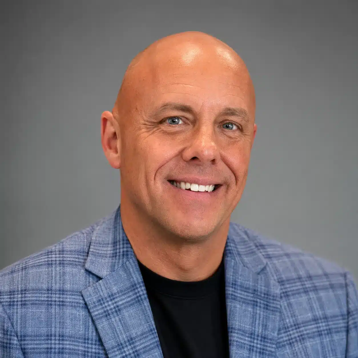 Smiling middle-aged man with a shaved head, wearing a blue plaid blazer over a black shirt, posed against a plain gray background.
