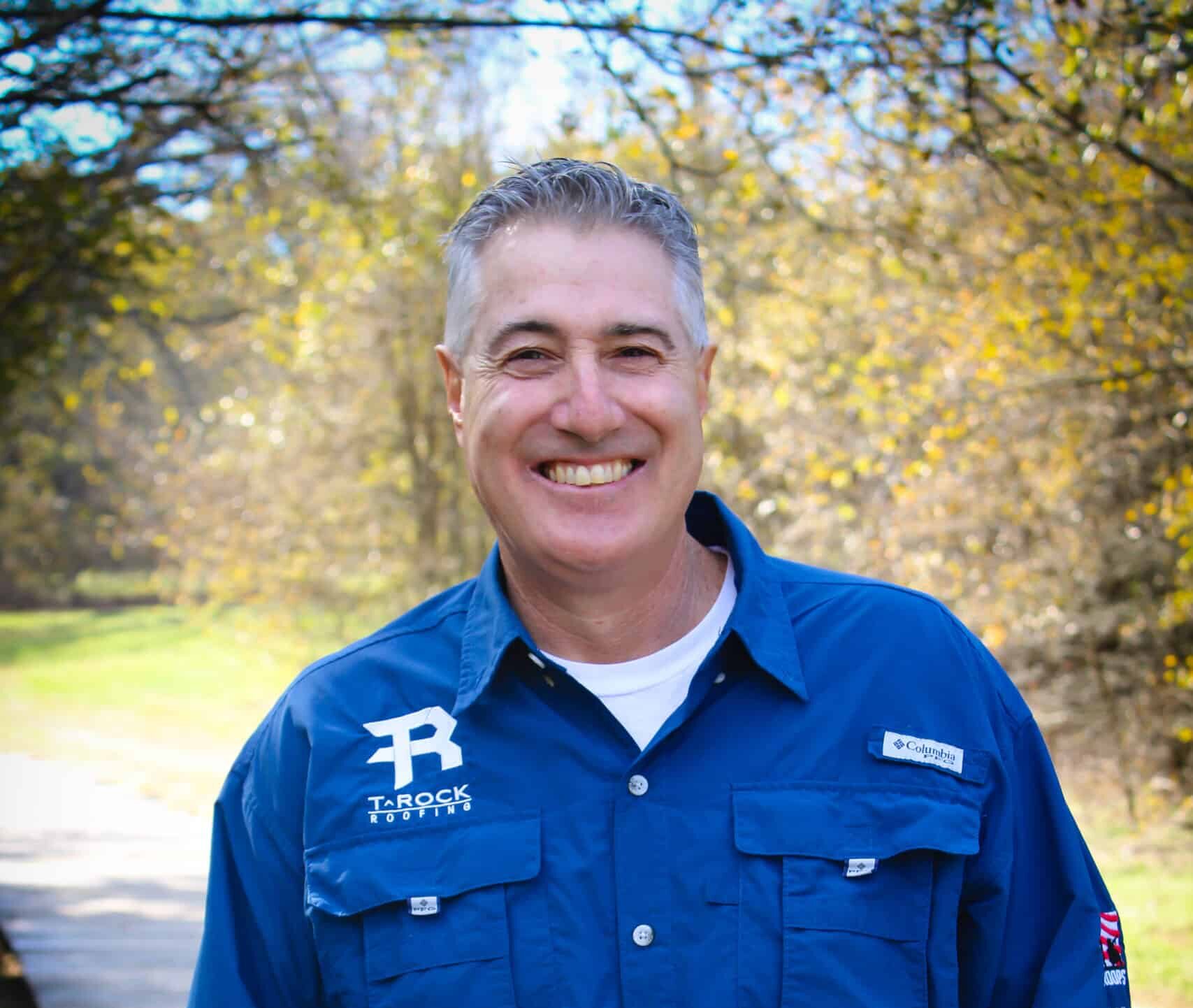 A smiling man stands outdoors on a wooden path surrounded by trees. He is wearing a blue button-up shirt with patches on the chest and sleeves, and has his hands in his pockets. The background is sunlit and features a walkway and greenery.