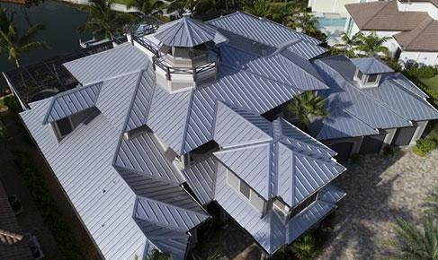 Aerial view of a large, modern house with multiple pitched metal roofs and a central tower. The house is surrounded by greenery and located near a water body, visible in the background. Paths and driveways made of stone or concrete are seen around the house.