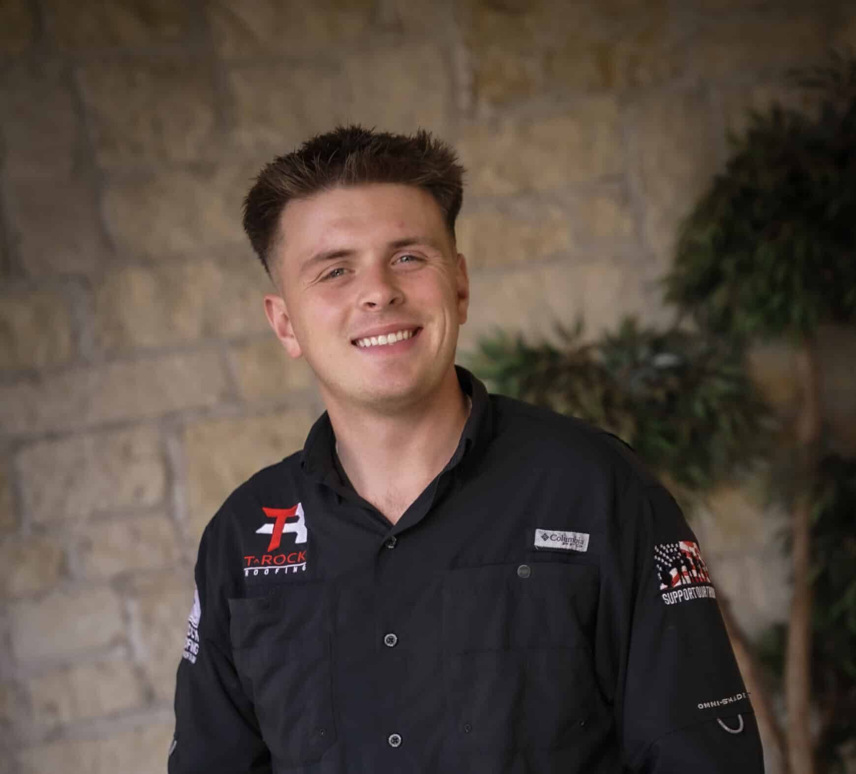 A young man with short brown hair smiles while standing with his hands in his jeans pockets. He wears a black button-up shirt with logos, a brown belt, and stands in front of a stone wall and green plant.
