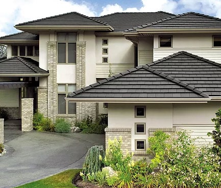 A modern two-story house with a light-colored exterior and dark gray tiled roof. The home features large windows, geometric design elements, and landscaped surroundings, including bushes and flowering plants near the entrance and driveway.