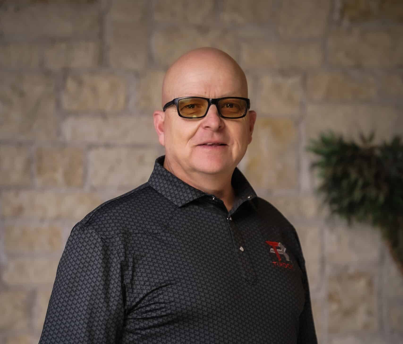 A man wearing glasses and a black patterned polo shirt stands in front of a stone wall, smiling slightly with his hands resting on his belt.