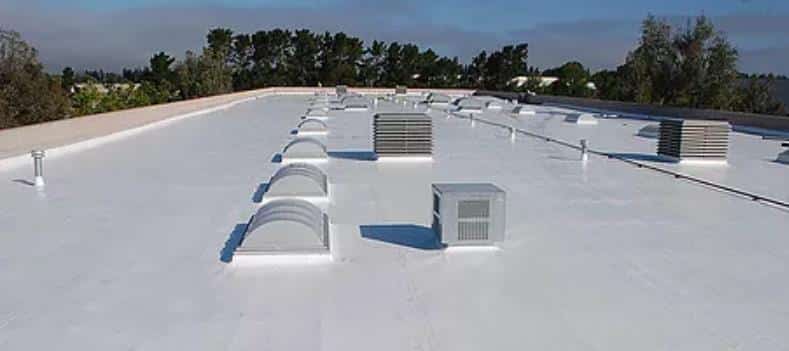 A large flat white rooftop with various HVAC units and vents spaced out across its surface. Trees and other buildings are visible in the background under a clear blue sky.