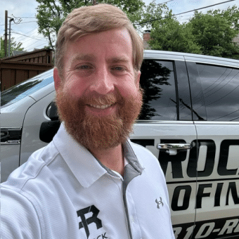 A man with a reddish-brown beard smiles for a selfie in front of a vehicle with the word "ROOFING" and a company logo visible on its side. He is wearing a white polo shirt with logos on it. Trees and a fence are in the background.
