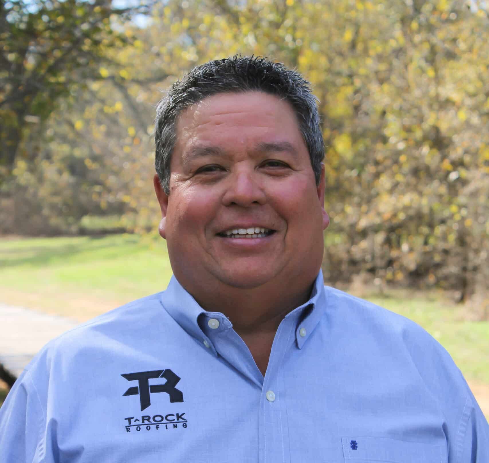 A man wearing a light blue button-up shirt with a "T-Rock" logo is smiling outdoors. The background features trees with brown and green foliage, suggesting a park or natural setting.