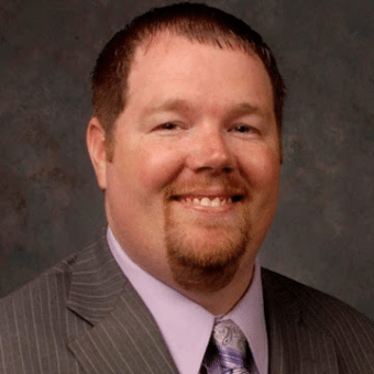 A man with short brown hair and a goatee is smiling. He is wearing a gray pinstripe suit, a lavender shirt, and a patterned tie. The background is a textured gray.