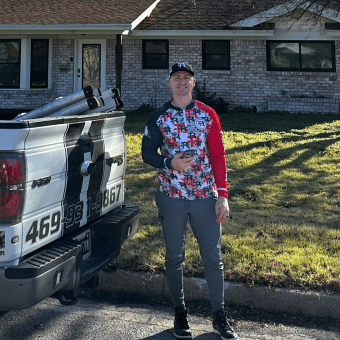A man in athletic clothing stands on grass next to a white pickup truck with black stripes and a phone number, in front of a brick house on a sunny day.