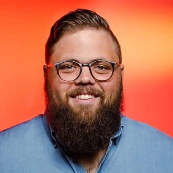 A smiling man with glasses, a full beard, and short, styled hair wears a light blue button-up shirt. The background features a gradient of orange and red tones.