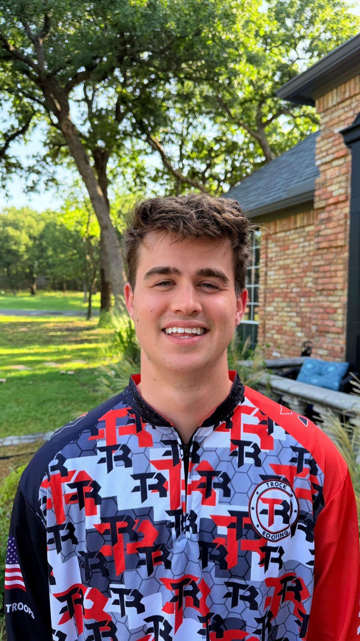 A young man with short brown hair smiles outside in a yard. He wears a colorful patterned shirt with “TR” logos and American flag on the sleeve. Trees and a brick house are visible in the background.