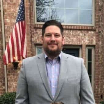 A man wearing a light gray suit and checked shirt stands in front of a brick house with an American flag and large windows visible in the background.