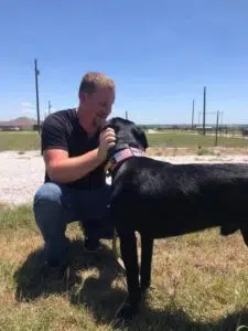 A man wearing a black t-shirt and blue jeans kneels on a grassy field, affectionately petting a black dog wearing a pink collar. The background shows a fenced area and clear skies above. - T Rock Roofing and Contracting A man wearing a black t-shirt and blue jeans kneels on a grassy field, affectionately petting a black dog wearing a pink collar. The background shows a fenced area and clear skies above.