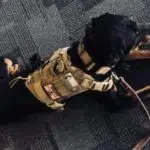 A black and tan dog wearing a tactical vest lies on a gray carpeted floor, gazing out through a window. The dog appears calm and attentive, with a leash attached to its vest.