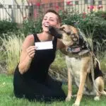 A woman kneels on the grass, holding a check, while a German Shepherd dog licks her face. The dog, adorned with a camouflage harness that has an American flag patch, sits beside her. They are surrounded by bushes and red flowers. Both appear happy.