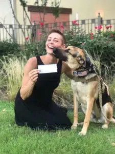 A woman kneels on the grass, holding a check, while a German Shepherd dog licks her face. The dog, adorned with a camouflage harness that has an American flag patch, sits beside her. They are surrounded by bushes and red flowers. Both appear happy. - T Rock Roofing and Contracting A woman kneels on the grass, holding a check, while a German Shepherd dog licks her face. The dog, adorned with a camouflage harness that has an American flag patch, sits beside her. They are surrounded by bushes and red flowers. Both appear happy.
