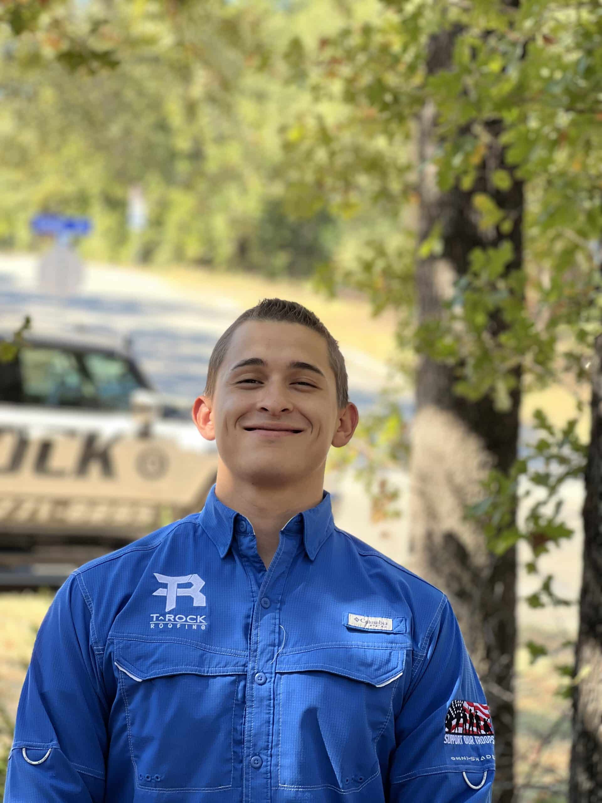 A young man with short brown hair wearing a blue collared shirt stands outdoors smiling, with trees and a police vehicle blurred in the background.