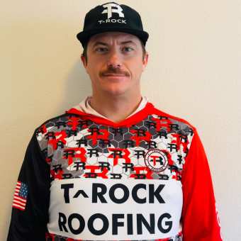 A person with a mustache wears a black T-Rock hat and a colorful T-Rock Roofing jersey with the American flag on the sleeve, standing against a plain light-colored wall.