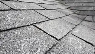 Close-up view of a grey asphalt shingle roof with circles drawn in white chalk, indicating areas of potential damage or inspection points.