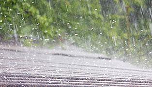 Heavy rain pours down, striking a surface and creating a splashing effect. In the blurred background, greenery is partially visible. The image captures the intensity and motion of the rainfall.