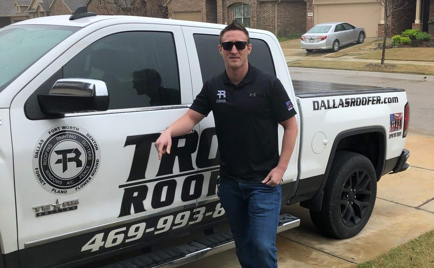 A man in sunglasses and black clothing leans against a white pickup truck branded with "Trox Roof" and "DallasRoofer.com." He stands on a residential street with houses and lawns in the background. The truck displays various logos and a phone number.