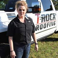 A woman with blonde hair stands in front of a white truck with the "T-ROCK ROOFING" logo on its side. She is wearing a black shirt and blue jeans. The background shows a grassy area and trees.