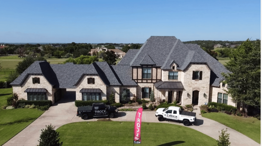A large, two-story stone house with a gray shingle roof is set against a suburban backdrop with plenty of greenery. Two work trucks are parked in the driveway next to a pink flag with the word "TROCK". The house has multiple windows and a well-manicured lawn.