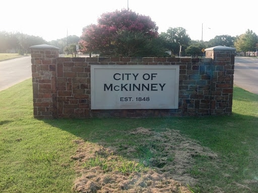 A decorative stone sign reads "City of McKinney, EST. 1848" and is surrounded by grass. The background features a tree with pink flowers and a road.