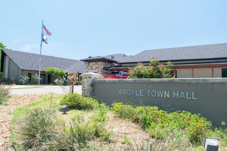 A sunny exterior view of Argyle Town Hall with a well-maintained garden. The building has a modern design with a stone and wood facade. An American and Texas flag stand on poles near the entrance. A grey-brick sign in the foreground reads "Argyle Town Hall.