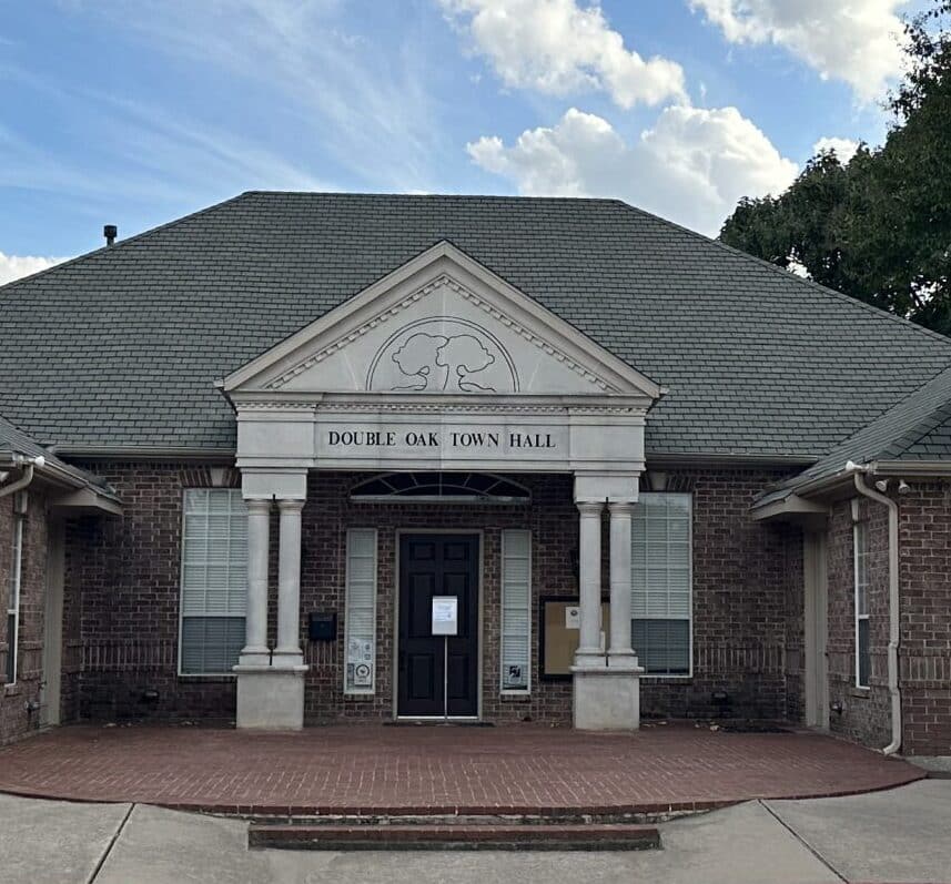 A two-story brick building with a gabled roof labeled "Double Oak Town Hall" above the entrance. The building features four white columns supporting a triangular pediment. There are two windows on each side of the black front door, and the driveway is in front.