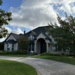 A large, gray stone house with tall, peaked roofs set against a partly cloudy sky. The house is surrounded by well-maintained green lawns and trees, with a curved driveway leading to the front entrance. Bright sunlight casts shadows, enhancing the texture of the stone facade.