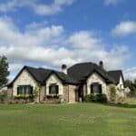 A stone house with dark green shutters and a black roof sits on a large, well-manicured lawn. Two trees stand on either side of the house. The sky above is blue with scattered clouds. Flower boxes with yellow blooms adorn the front windows.